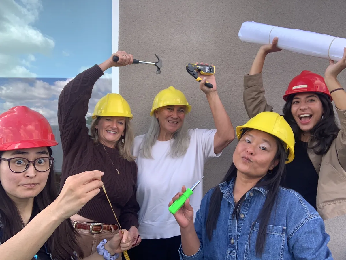 Five team members in hard hats striking poses with a hammer, drill, tape measure, and rolled-up plans