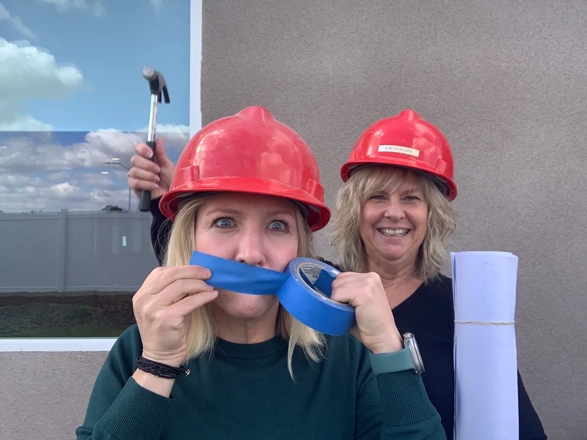A team member in a red hard hat with painter's tape over her mouth while a coworker laughs