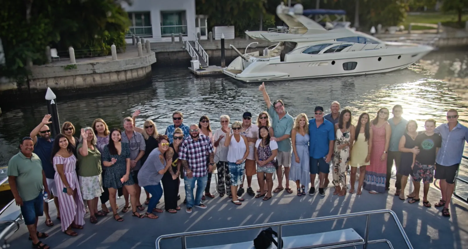 About thirty S&S Homes employees gathered on a boat deck, waving, with a yacht in the harbor behind them