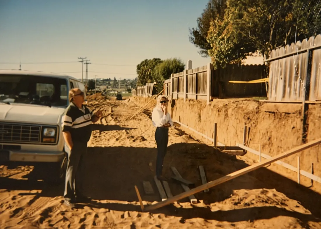 Two people walking a freshly graded job site beside an open foundation trench