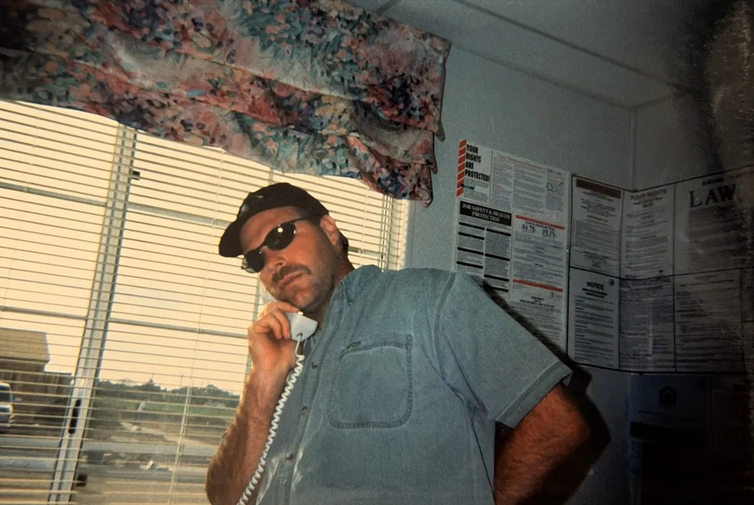 Steve Sanders on a corded phone in a construction office, wearing sunglasses and a cap